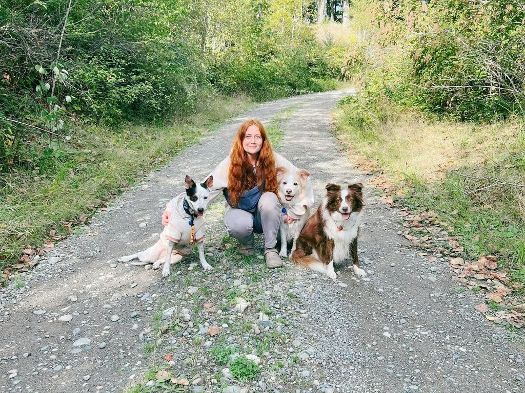 Marta crouching on a trail with three dogs — two white dogs and a red-and-white border collie — sitting calmly beside her